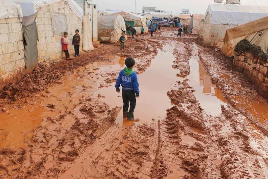 Group of children standing on dirty wet ground with puddles between old tents in refugee camp with i