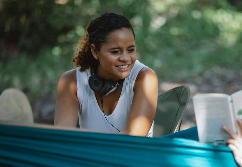 Happy young Hispanic female with headphones sitting on chair near friend in hammock with book while 